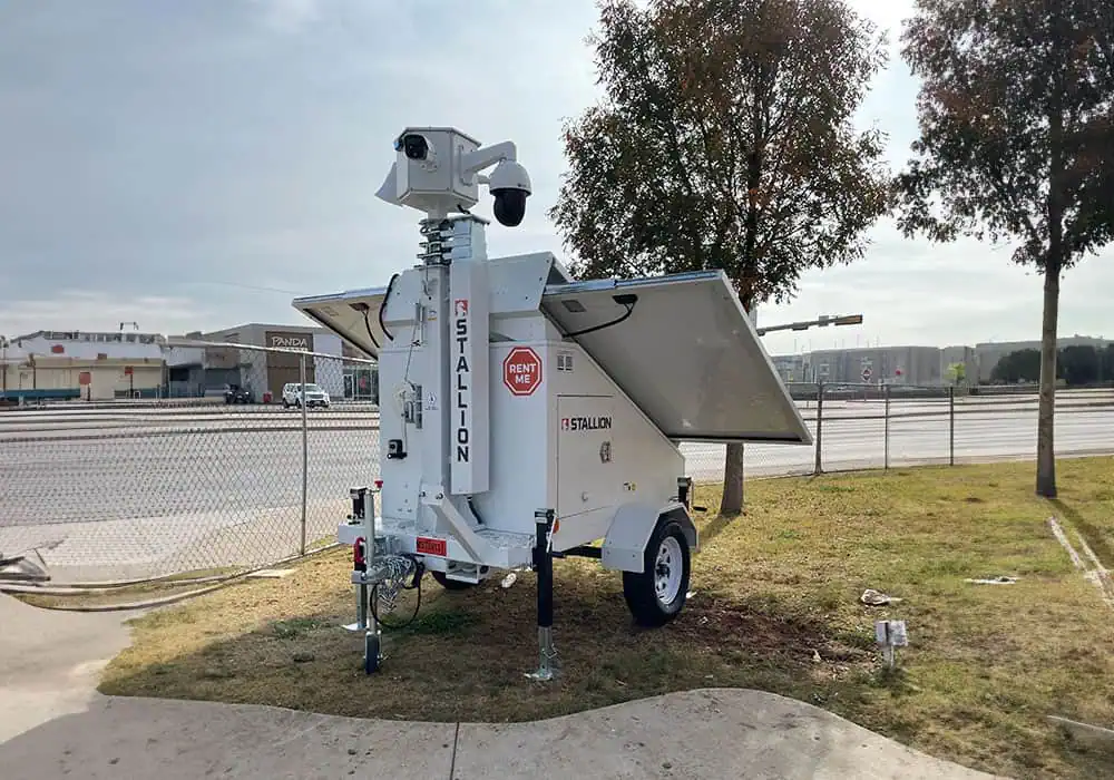 A mobile surveillance trailer with solar security cameras provides parking lot and commercial video security near a fenced roadside.