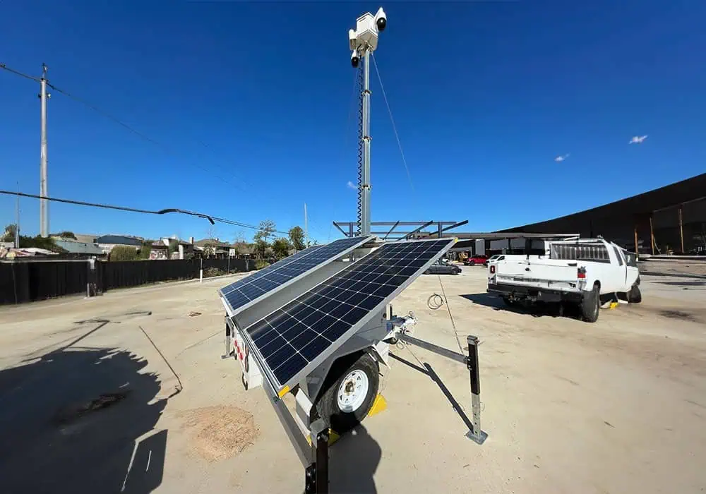 A mobile surveillance trailer with solar security camera provides parking lot security in an industrial area near a white pickup and commercial buildings.