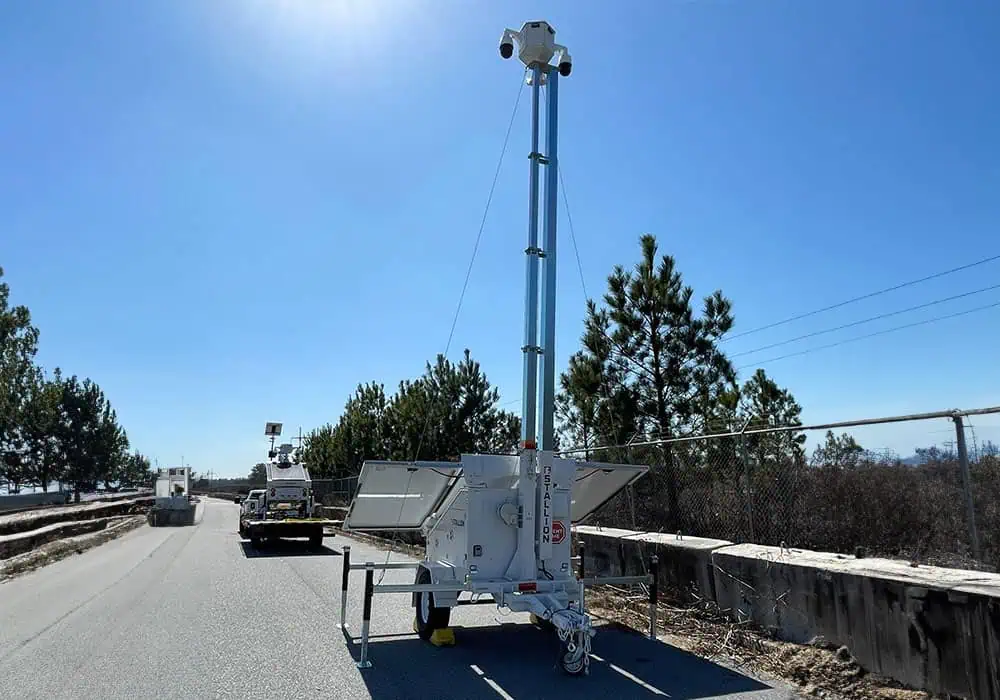 A mobile surveillance trailer with solar security camera offers parking lot security by a chain-link fence under a blue sky.