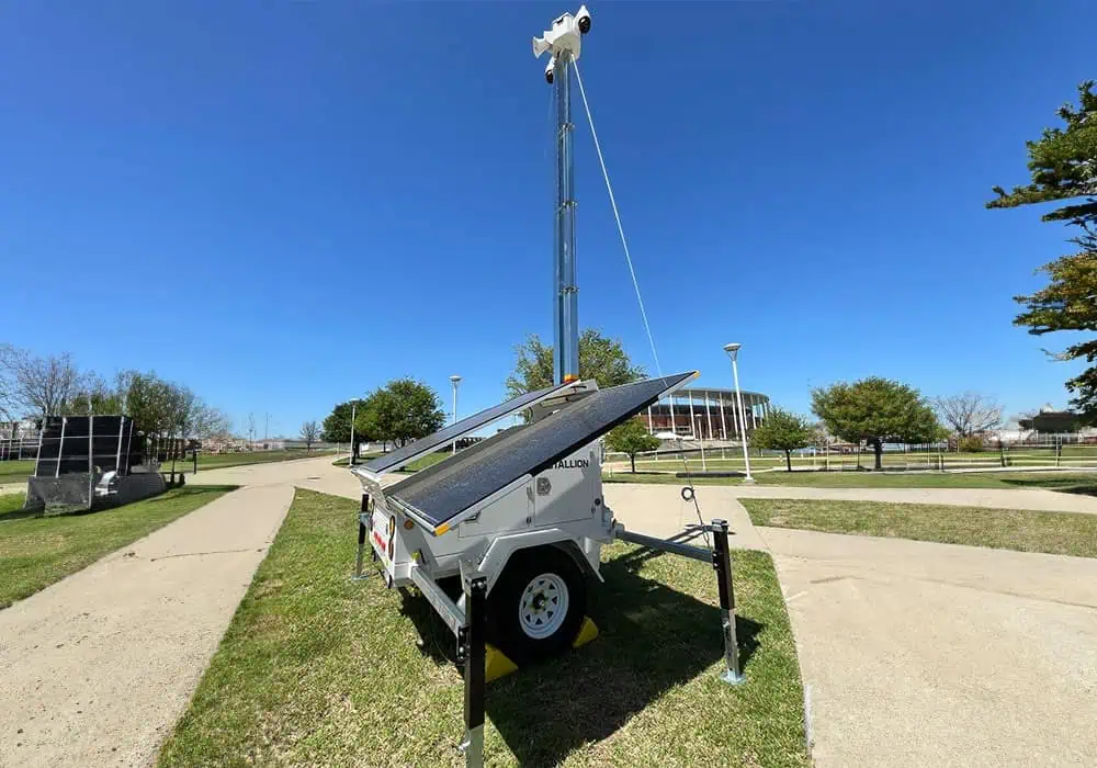 A mobile surveillance trailer with solar security camera provides parking lot security on grass near walkways under a clear blue sky.
