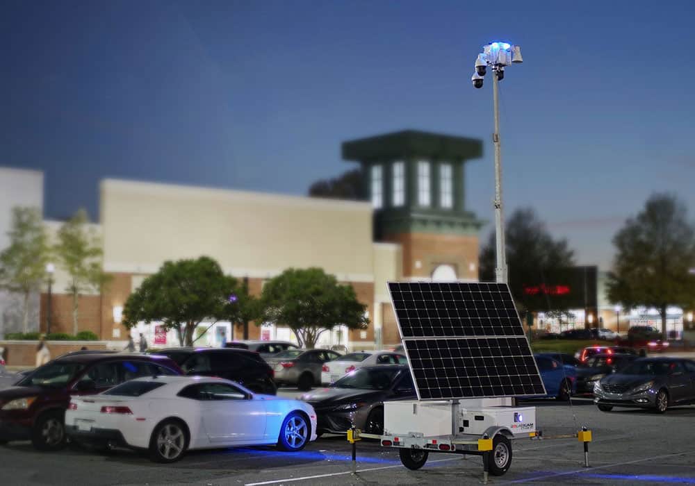 A mobile surveillance trailer with solar security camera provides parking lot security near a shopping center at dusk.