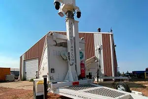 A mobile surveillance trailer with solar security cameras is parked by a metal building for commercial parking lot security.