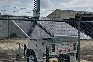 A solar-powered mobile surveillance trailer with solar security camera and raised antenna is parked behind a fence, suited for parking lot security.