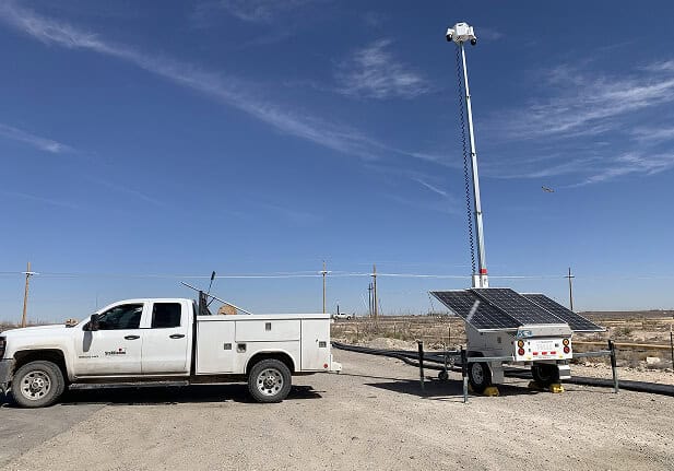 A white utility truck beside a mobile surveillance trailer with solar security cameras and a tall commercial security mast for parking lot security.